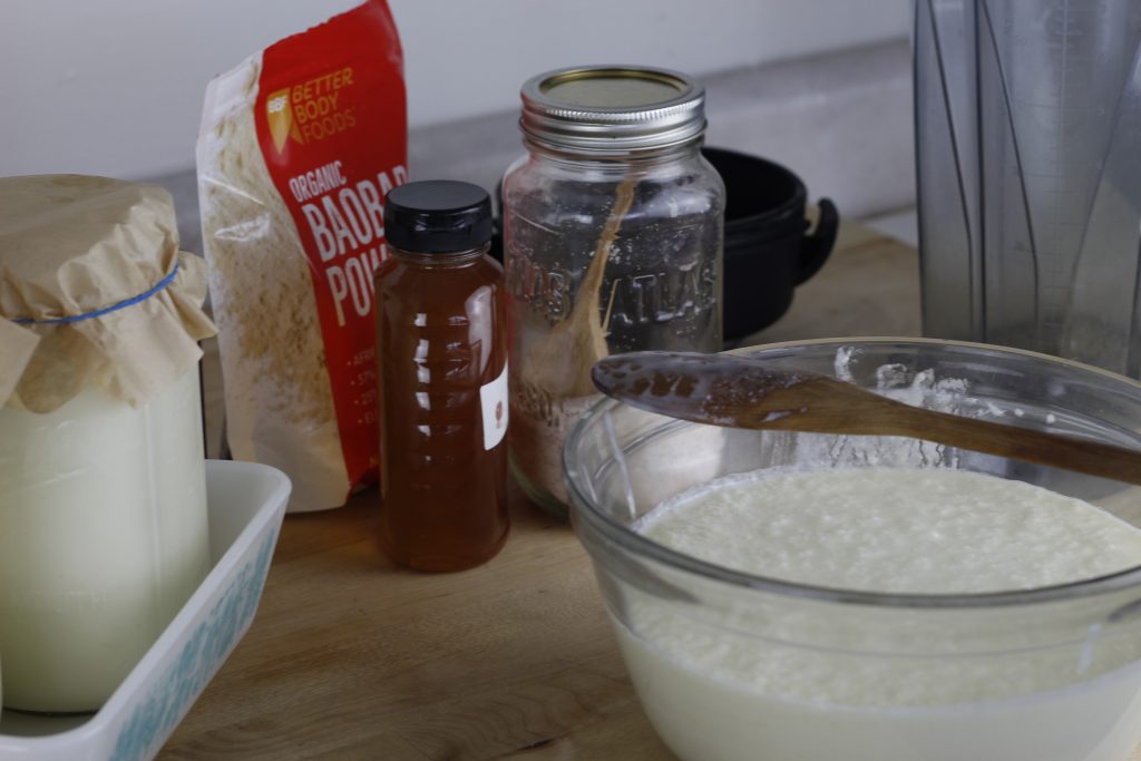 image of ingredients for a healthy chocolate kefir smoothie recipe: honey, baobab powder, salt kefir cocoa powder, on a counter top 