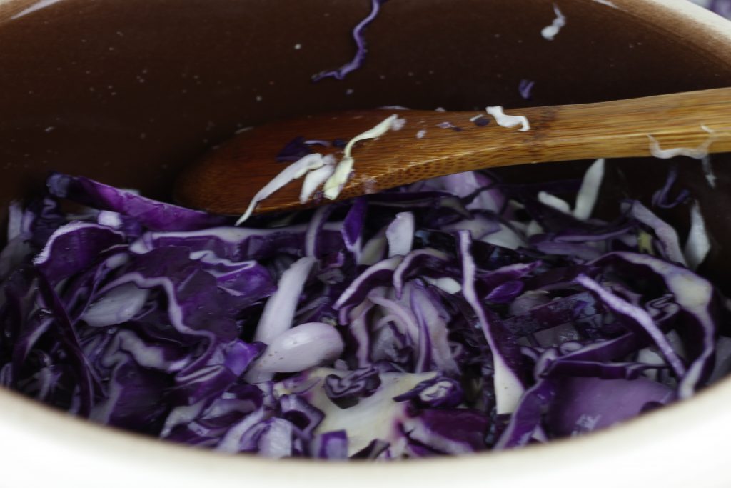 image of purple cabbage in a fermenting crock being stirred with a wooden spoon