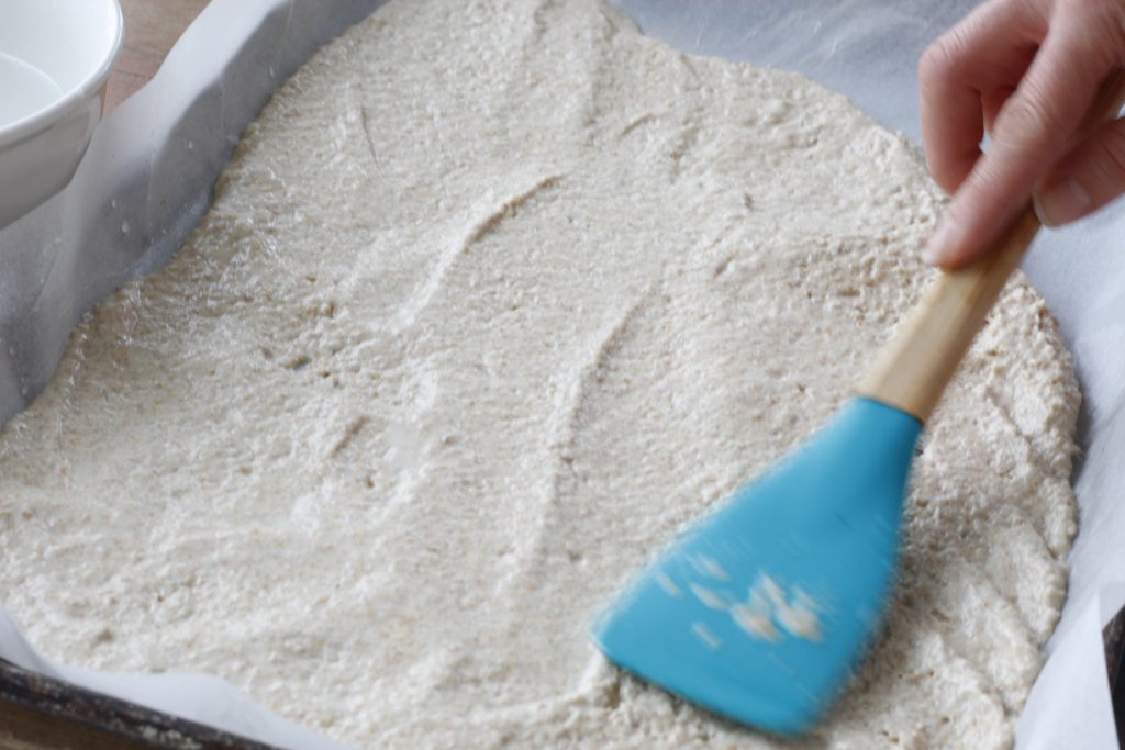 image of rice and sourdough discard cracker dough being spread to the edges of the baking sheet with a blue spatula