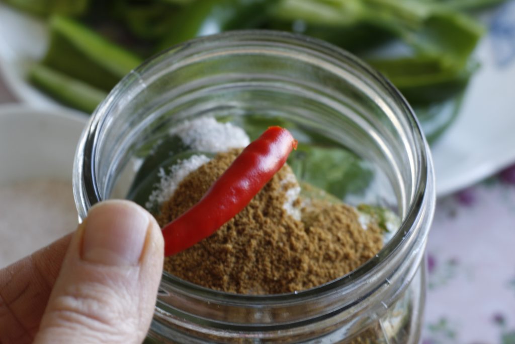 close up image of a hand holding a hot red pepper above a jar and spices measured on the top 