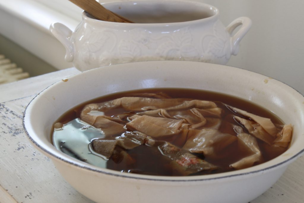 image of fabric soaking in a tea-dye bath in a bowl on a white table behind it is another dish with a wooden spoon resting in it