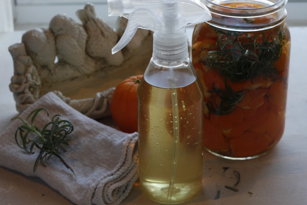 image of orange-rosemary cleaner in a clear spray bottle in front of an orange and beside a cleaning cloth with a rosemary sprig and a jar of cleaner 