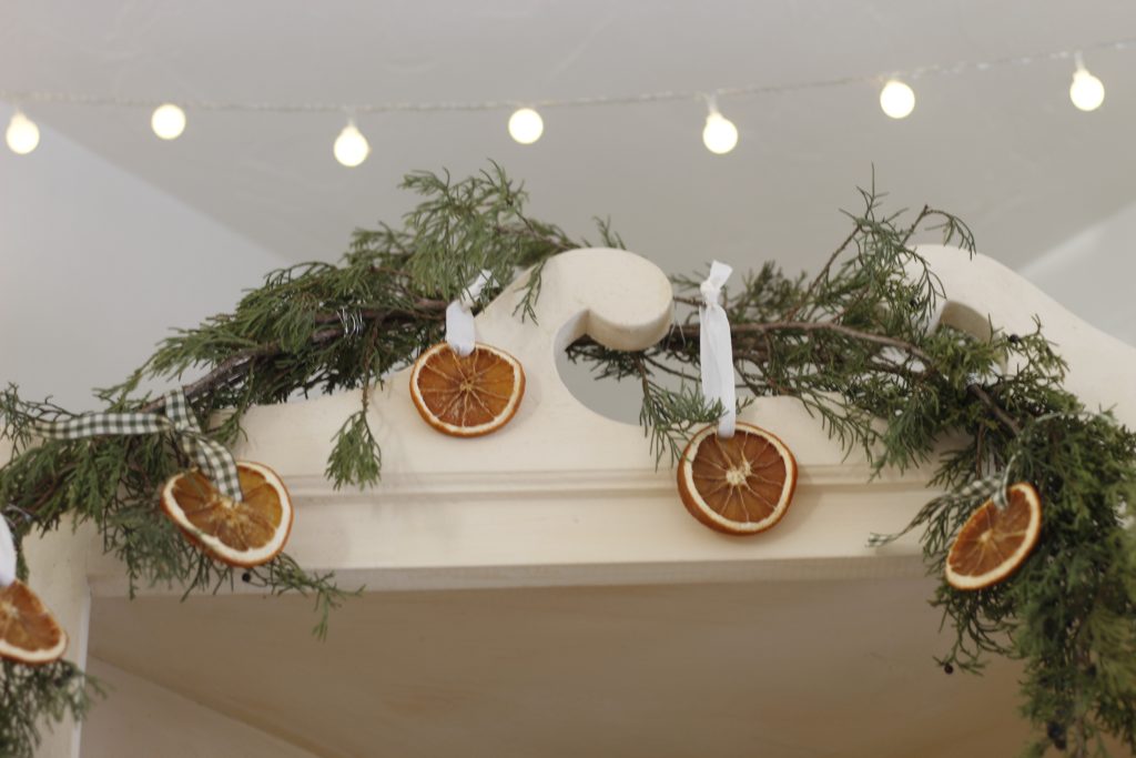 Image of a garland of cedar and dried orange slices draped across the top of a cream colored cabinet and strand of lights hung above it