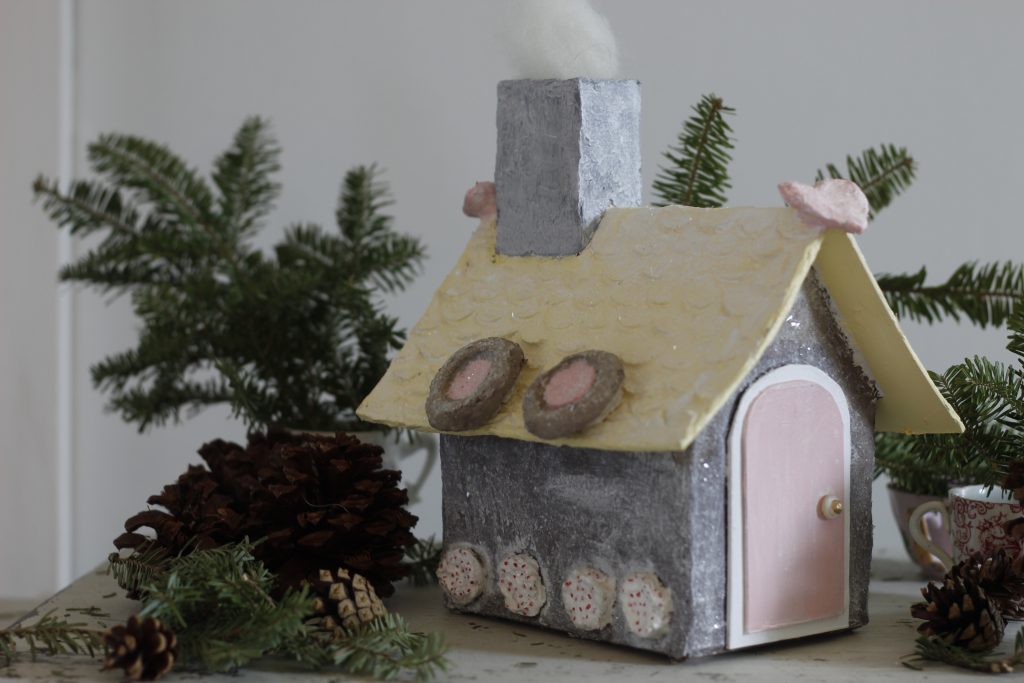 image of papier-mache house on a table with greenery and pinecones around it