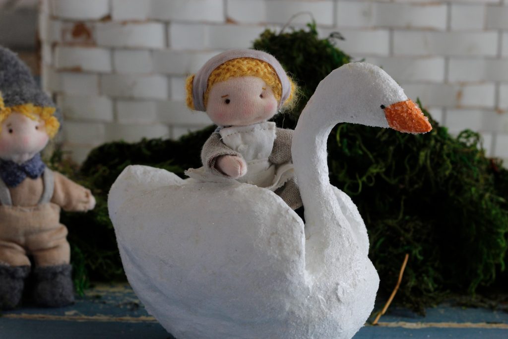 image of a white papier-mache duck with a small handsewn girl doll riding on its back in the background is a small handsewn boy doll standing in front of a white background and green moss 