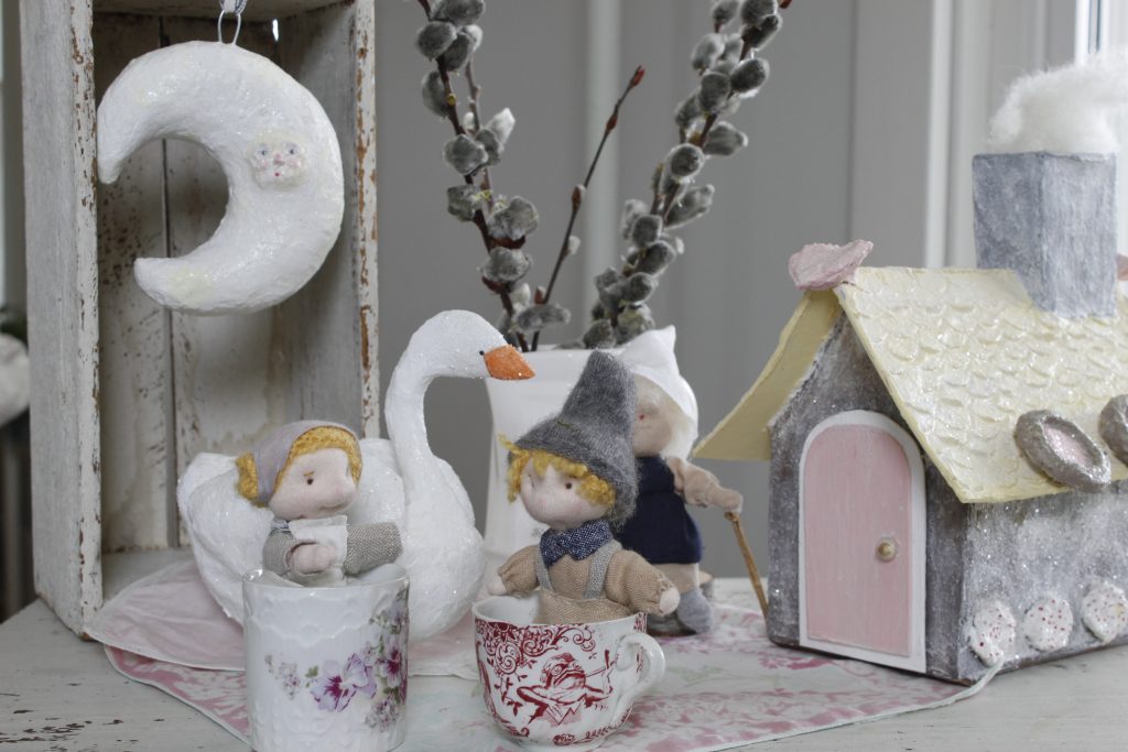 image of handsewn fabric Hansel and Gretel dolls resting inside tiny pretty teacups and in the background is a white papier-mache duck and a hanging papier-mache moon and a pitcher of cut plants and a handsewn fabric witch doll standing by her papier-mache gingerbread house