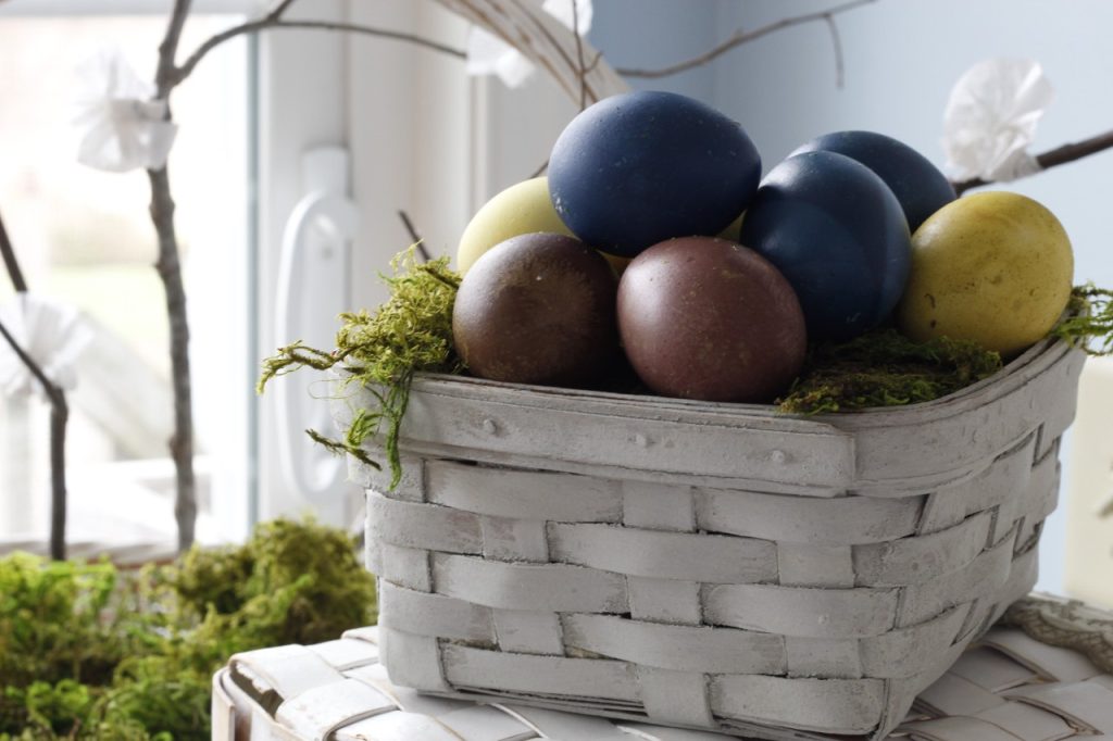 image showing a gray painted basket holding Easter eggs dyed with plant dyes in the foreground, there is green moss sticking out of the basket, in the background is another basket holding flowering branches and moss