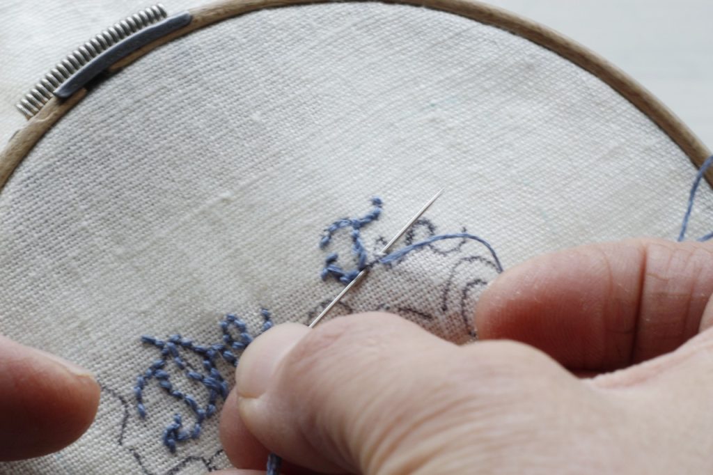 This is an image of hands embroidering a letter with blue thread on white fabric.