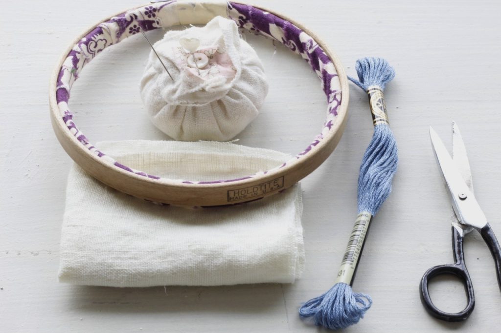 This is an image of embroidery supplies lying on a table they are fabric,, white pincushion, embroidery hoop, blue embroidery floss, a pair of black handled scissors