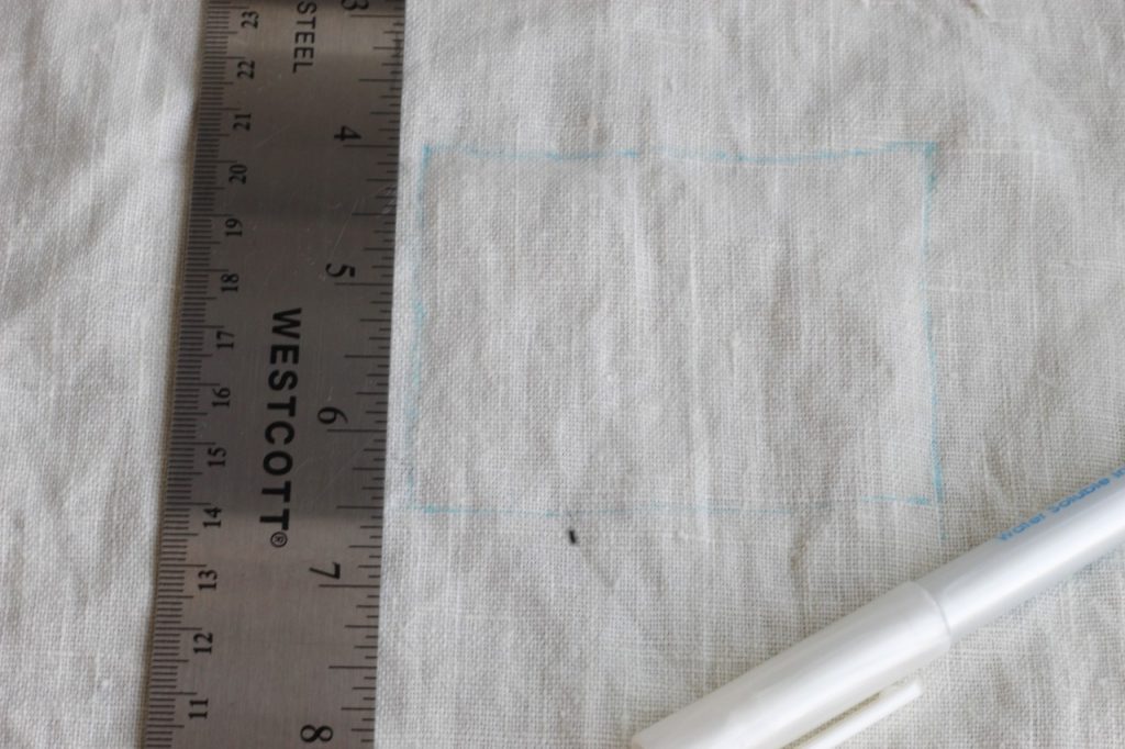 This is an image of linen fabric that has been marked with a blue rectangle for placement of an embroidery design and there is a metal ruler along the left side of the image