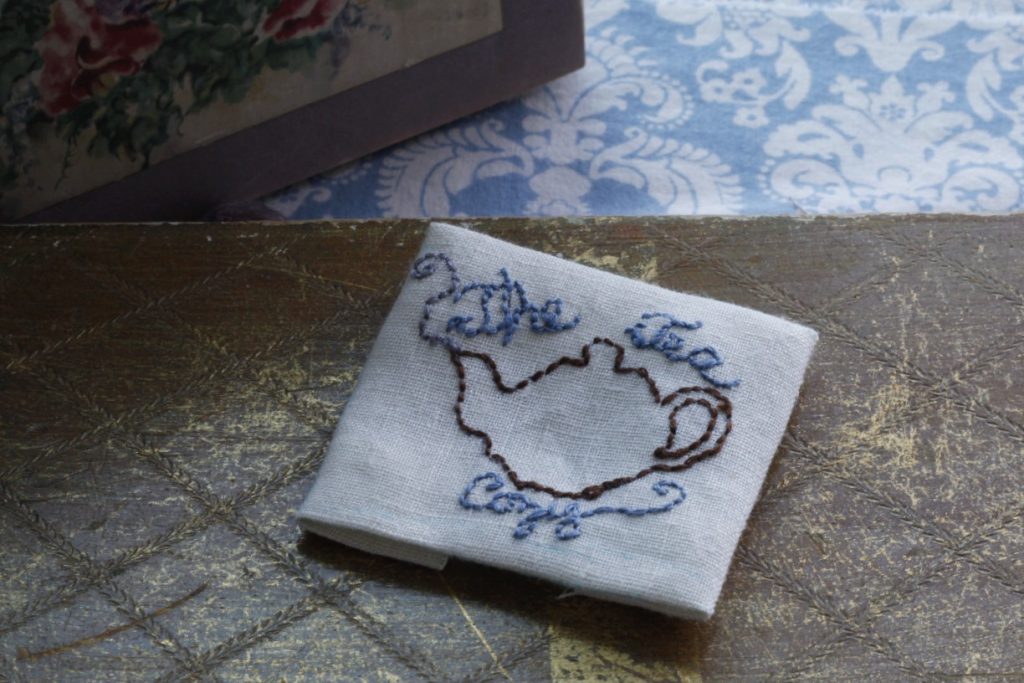 This is an image of a finished simple embroidery design on a golden box on a blue and white tablecloth and in the background is a floral purple decoration