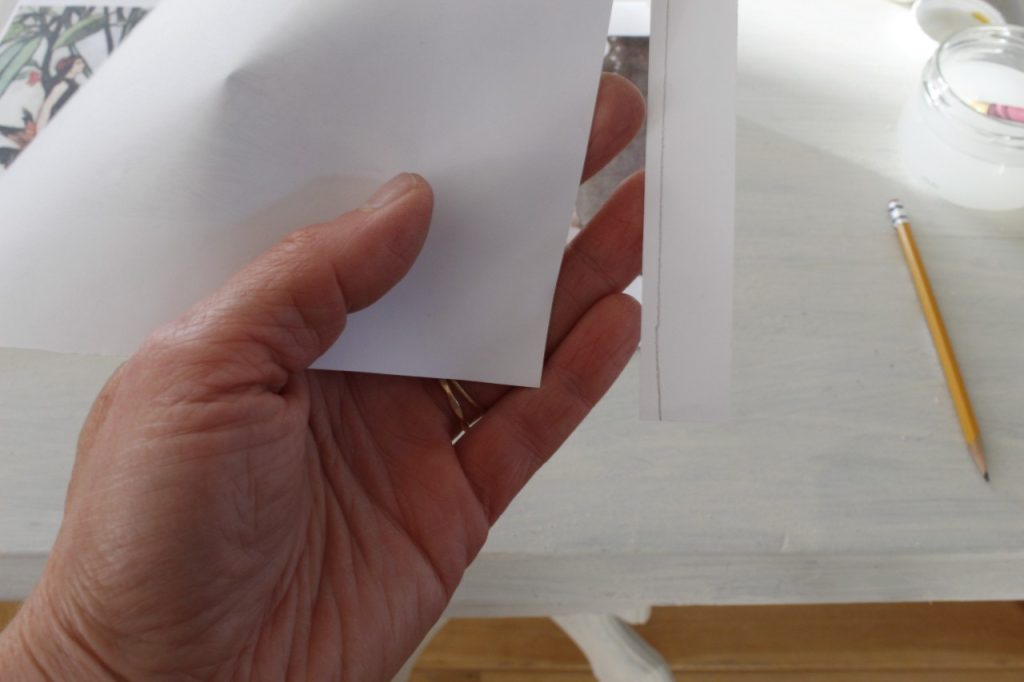 This is an image of a hand holding a paper with a traced line drawn on it and showing that it has been cut about 1/8 inch from the line. There is a white table and a pencil in the background.