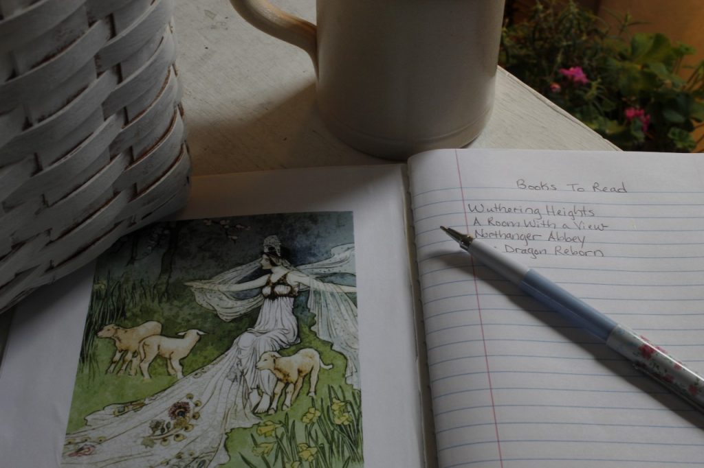 This is an image of a finished open notebook with a books to read list started. There is a pen lying on the book. In the background is a white basket, a beige pottery mug and a pot of flowers.