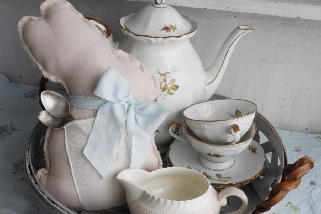 This image shows the mini bunny pillow on a basket as a tea tray holding a teapot and teacups, in front of all that is a pink and white creamer. The pocket on the pillow is holding two silver teaspoons.