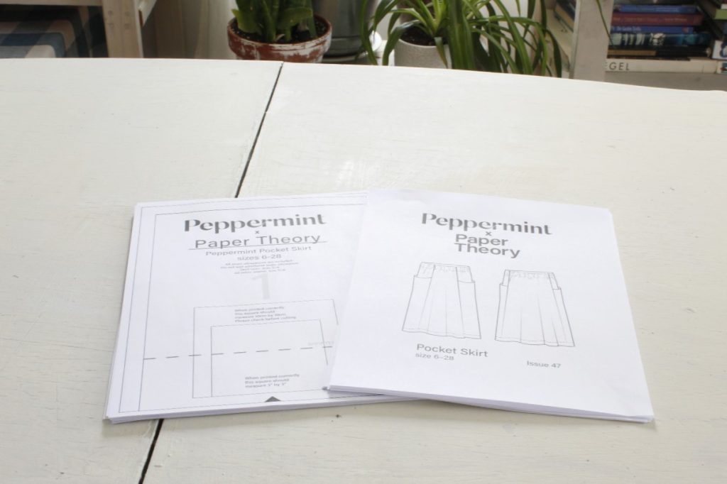 This is an image of a stack of printed out pattern pages on the left and papers in a stack on the right that is the instructions of a Peppermint Magazine sewing pattern of a pocket skirt, they are lying on a white table and in the background are books and houseplants.