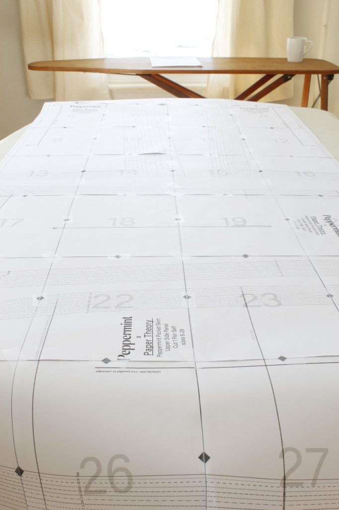 This is an image of the sheet of pattern pages laid out on a table and they have all been taped together. In the background is a window and cream-colored curtains and a wooden ironing board with a coffee mug set on top of it.