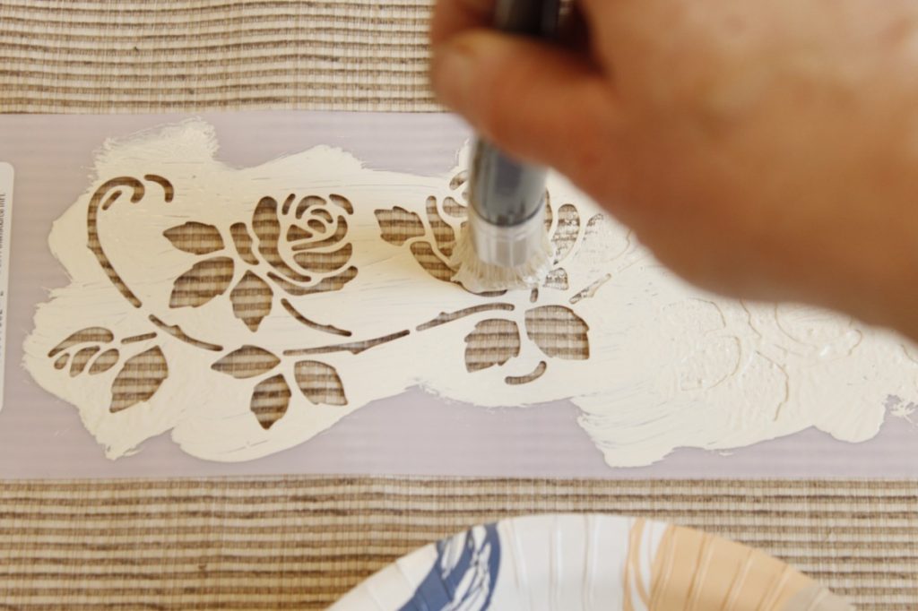This image shows a hand holding the stencil paintbrush and filling in the open areas with paint and holding the paintbrush vertically while tapping the paintbrush onto the fabric.
