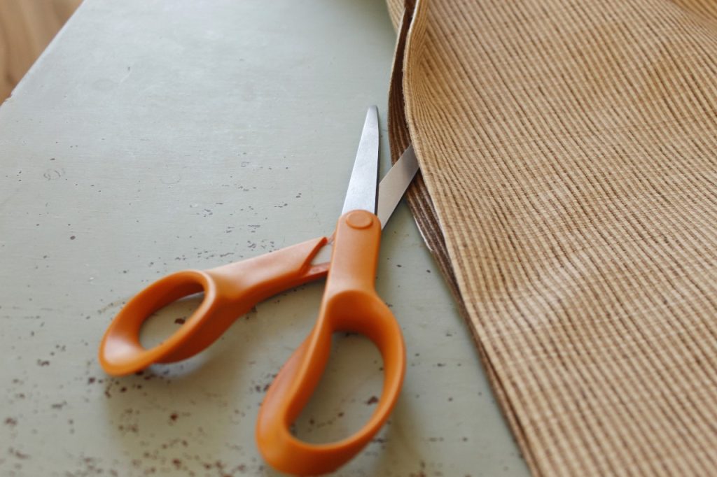 This image shows the beige fabric lying on a painted table being cut with orange scissors.