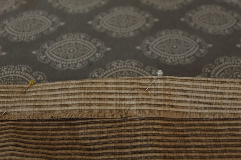 This image shows the beige upholstery fabric lying on a grey fabric covered ironing board with a hem folded over and pins holding it.