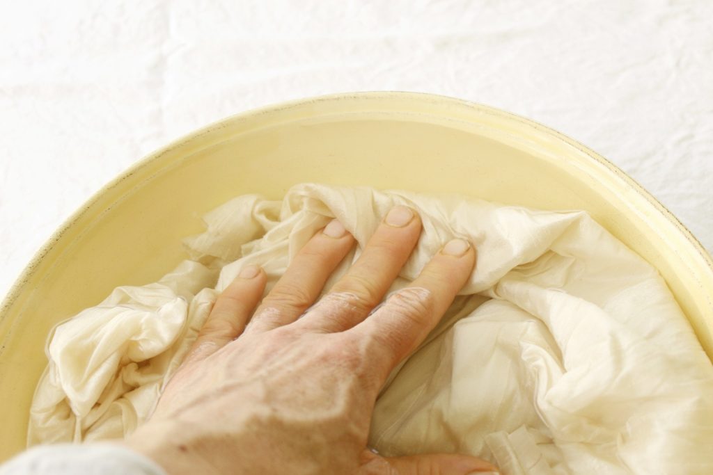 This image shows the ivory silk fabric being submerged in a yellow bowl of water by a hand.