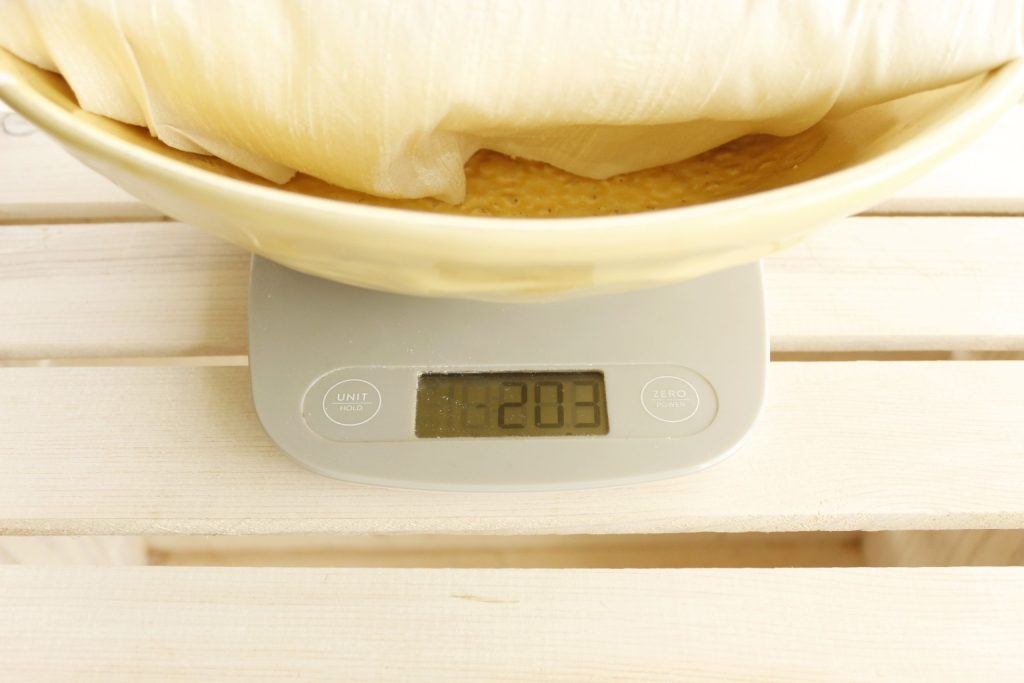 This image shows ivory colored silk fabric folded in a yellow bowl being weighed by a digital scale to calculate amount of mordant to use.