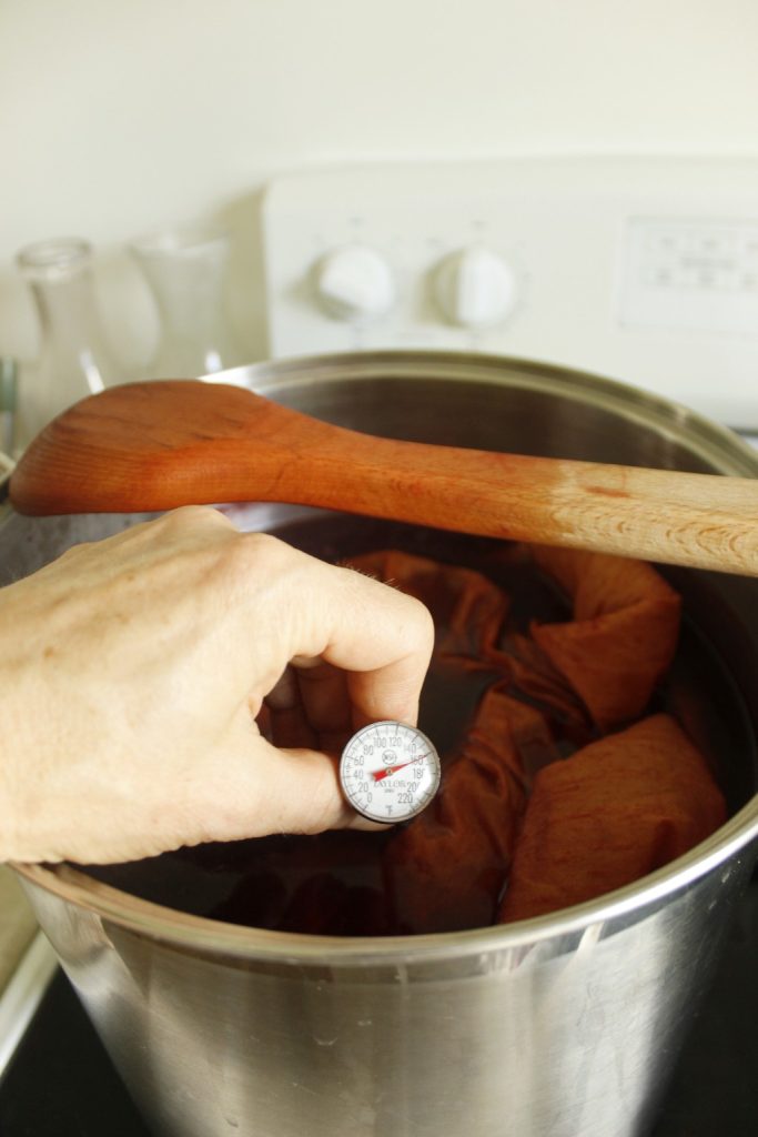 This image shows the silk fabric in the dye pot of madder root. There is a hand holding a thermometer in the pot to make sure the temperature stays relatively the same. There is a wooden spoon propped on the dye pot.