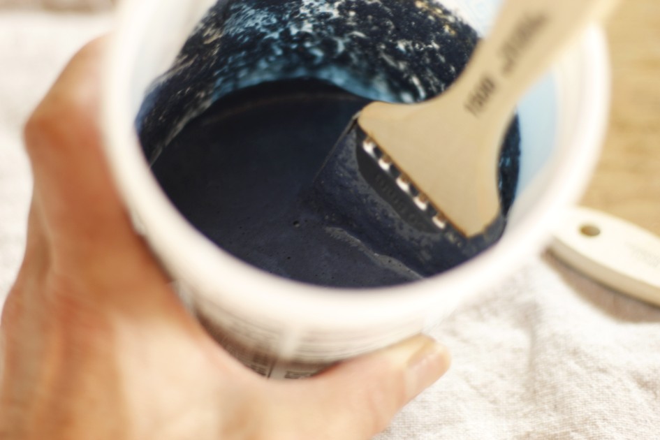 This image shows dark blue paint milk paint mixed with water in a recycled container with a paint brush , being held in a hand.