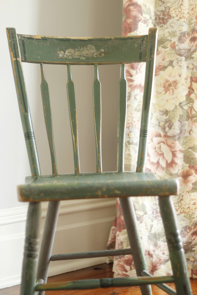 This is an image of an old chipped green dining chair that has been painted over with gray paint that is also chipping, setting by a window with a long floral curtain.